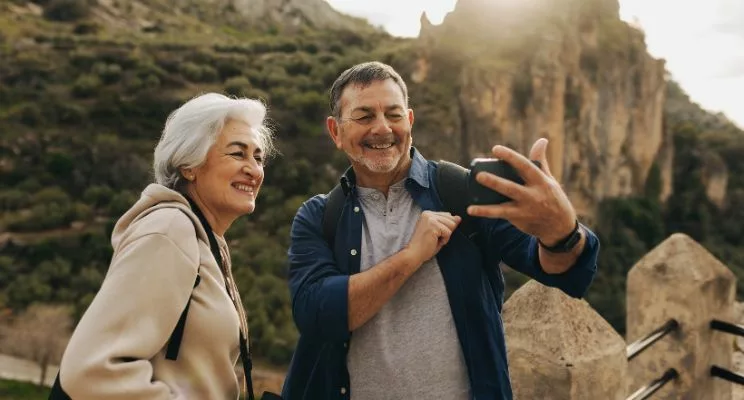 An older couple enjoying a moment outdoors, capturing a memory together with a smartphone camera. The couple stands side by side, smiling, as they use the phone to take a picture, surrounded by the beauty of nature.